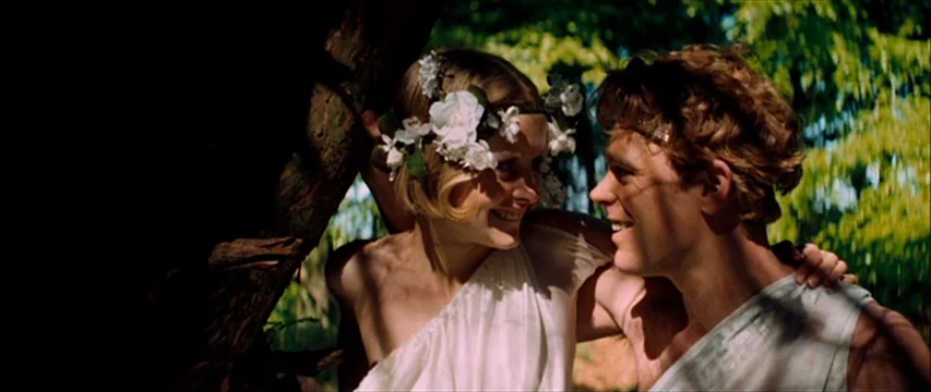 The Boy Friend (1971) movie still: medium — A sunlit outdoor area with dense leafy green foliage providing a background. A young couple wearing…