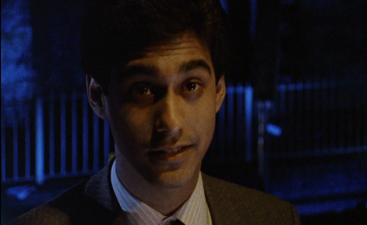 My Beautiful Laundrette (1985) movie still: closeup — An outdoor urban area at night with a metal fence visible in the background. A young man…