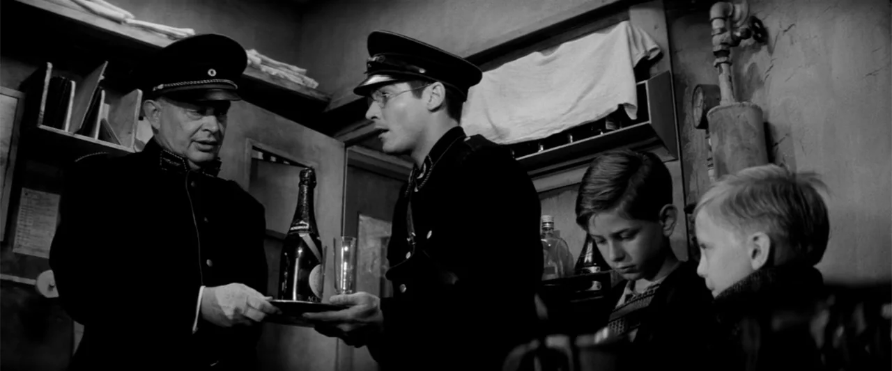 Europa (1991) movie still: medium — A cramped concrete bunker features exposed pipes and basic wooden shelving on the walls. Two uniformed men and…