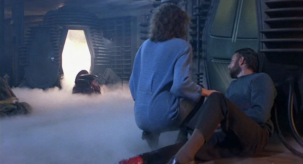 The Fly (1986) movie still: medium — A dark spaceship corridor filled with thick ground-level fog and large industrial machinery. A woman kneels…