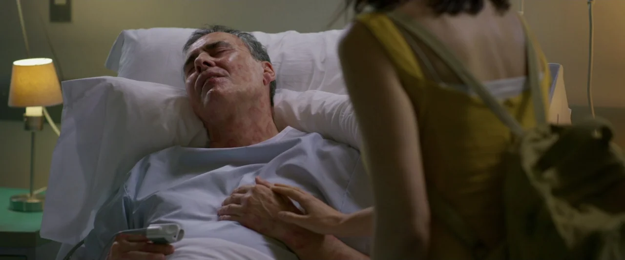 Dog Life (2024) movie still: medium — A hospital room contains a patient bed with white linens and a bedside lamp. A woman wearing a backpack stands…