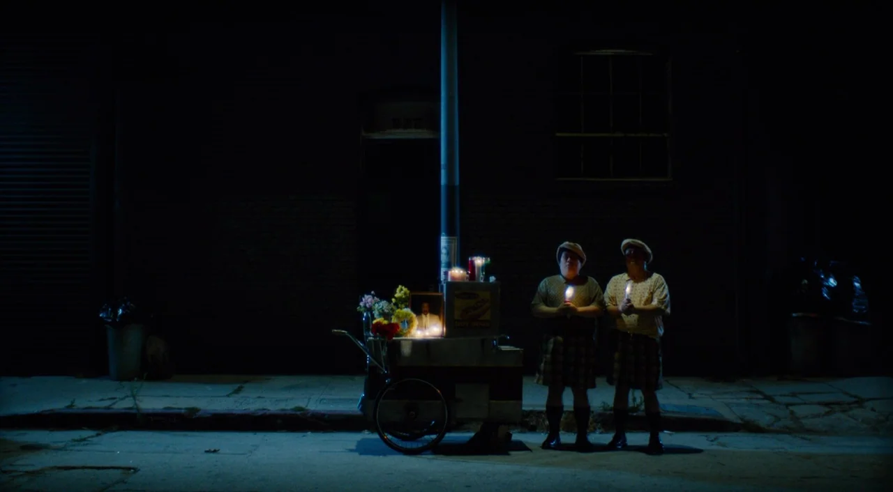 The Greasy Strangler (2016) movie still: wide — Two men stand on a dark city sidewalk at night beside a memorial cart. The memorial features…