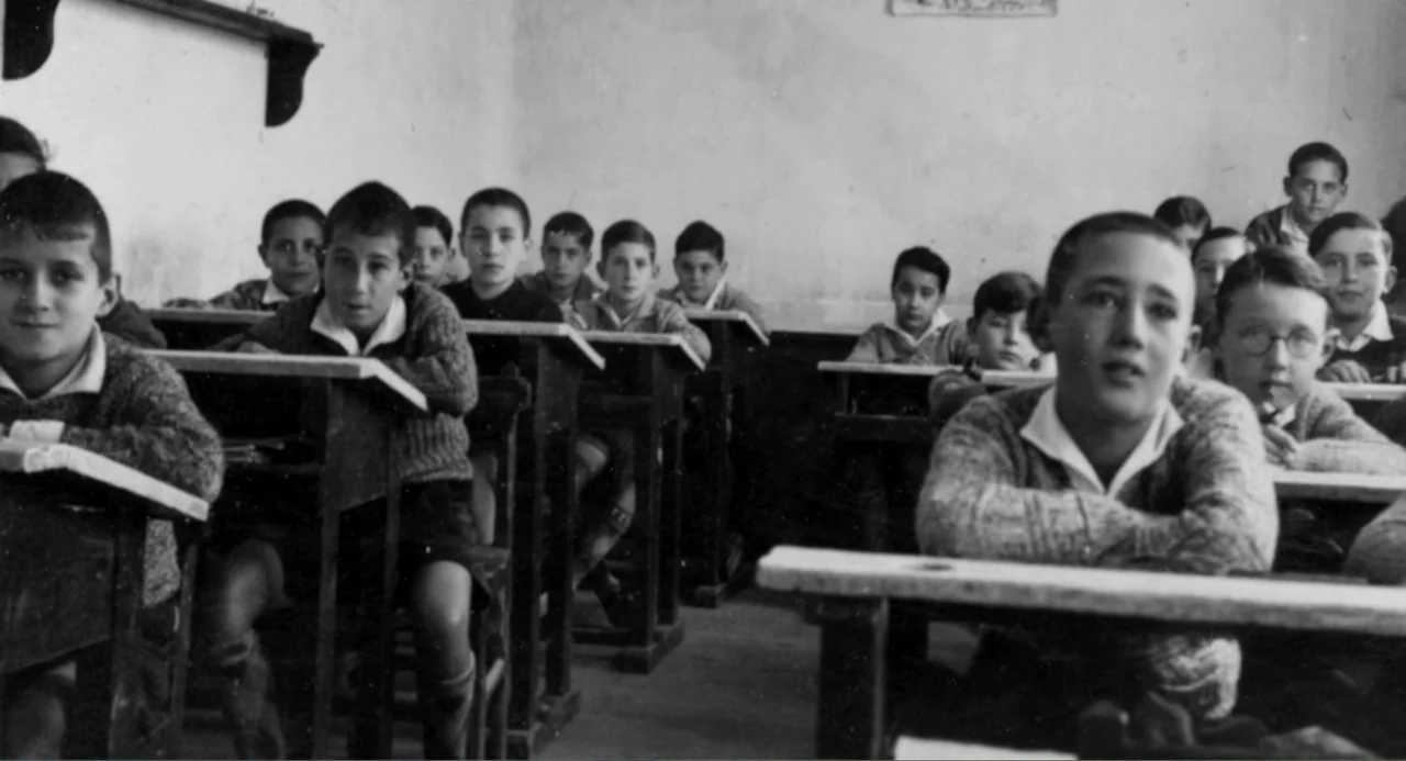 Los días azules (2025) movie still: medium — A vintage classroom with multiple rows of wooden desks and students seated together. Multiple young…
