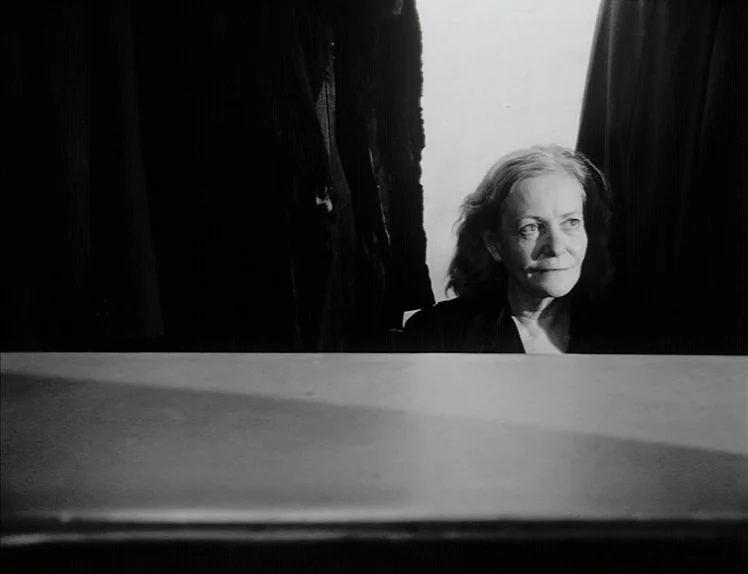 Damnation (1988) movie still: medium — A woman is positioned behind a large dark flat surface in an interior space. Heavy black curtains hang in the…