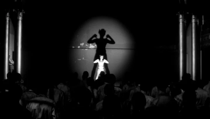 The American Astronaut (2001) movie still: wide — A dark theater with rows of obscured audience members facing a lit stage area. A performer stands…