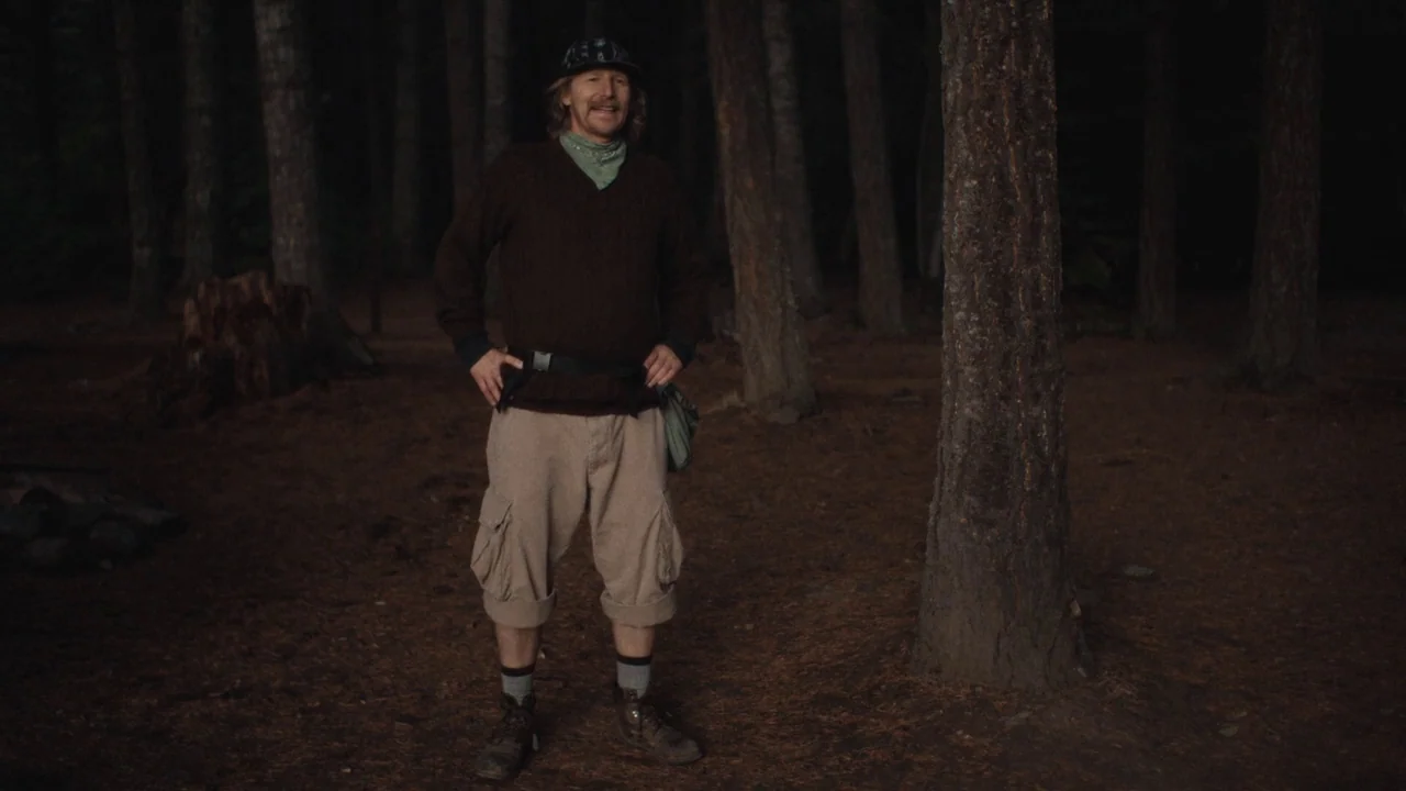 Night Moves (2013) movie still: fullbody — A man stands in a dark wooded area filled with tall pine trees. He stands still with his hands on his…