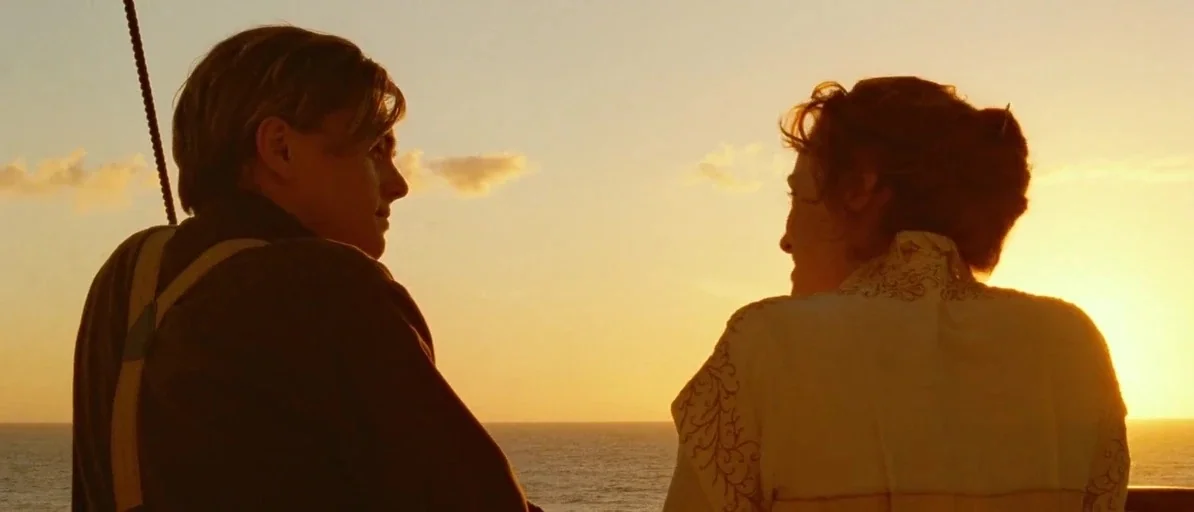Titanic (1997) movie still: medium — A man and a woman stand on the railing of a ship at sunset. They face each other in profile against a golden…