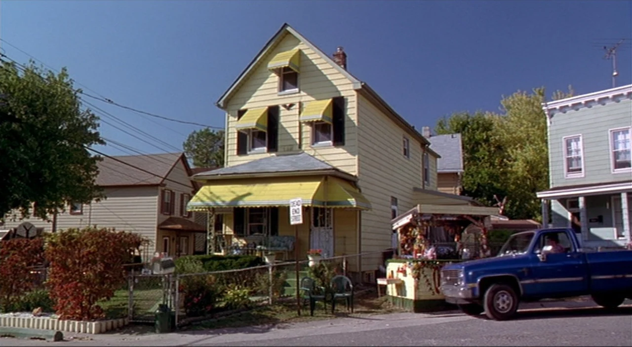 Pecker movie still: establishing — A light yellow wooden two-story house stands on a residential street with suburban homes. A blue pickup truck is…