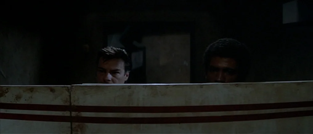 Assault on Precinct 13 (1976) movie still: medium — Two men hide behind a partition marked with two horizontal red stripes. The background is a…