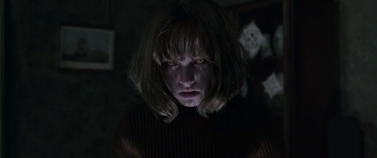 The Conjuring 2 (2016) movie still: closeup — A dimly lit domestic interior features walls with dark patterns and framed artwork. A young woman with…