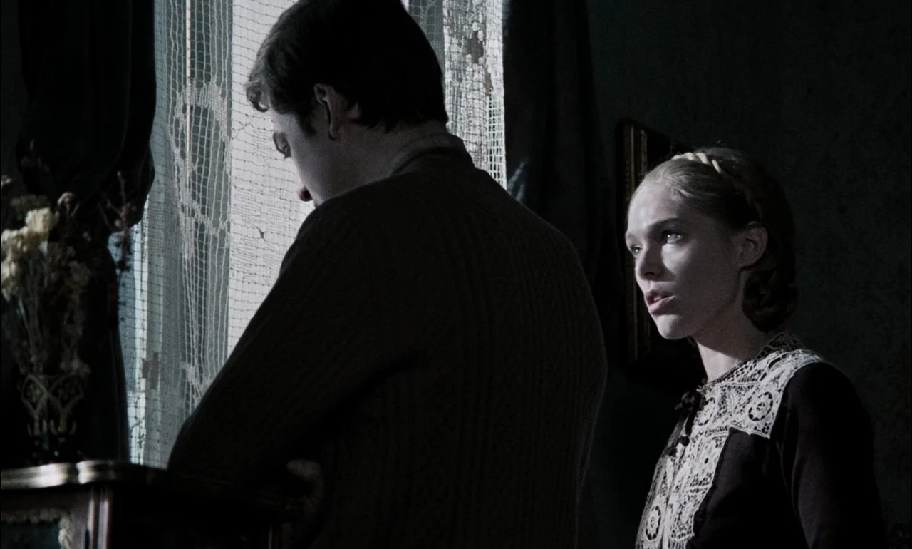 The Third Part of the Night (1971) movie still: medium — A dimly lit room features a man standing before a window with lace curtains. A young woman…