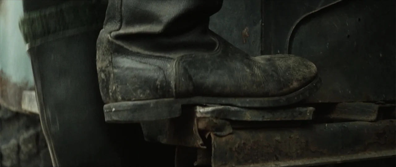 Sisu: Road to Revenge (2025) movie still: closeup — A close-up view of a worn, mud-caked leather boot resting on a metal step. The surface is…
