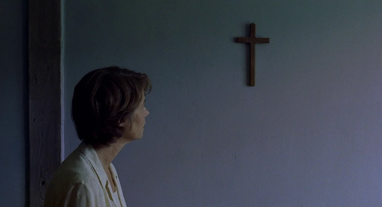 Swimming Pool (2003) movie still: medium — A plain, dimly lit room features a simple wooden cross attached to a wall. A woman in a light-colored…