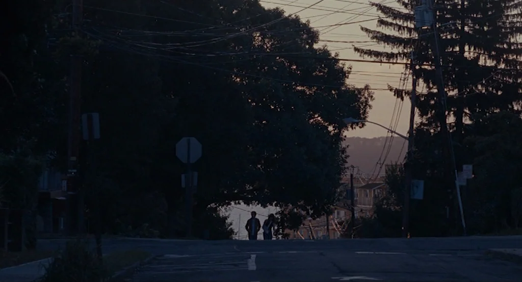 Louder Than Bombs (2015) movie still: wide — A quiet, paved residential street at dusk is flanked by dense, silhouetted trees. Two figures walk side…