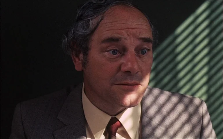 Business is Business (1971) movie still: closeup — An interior space features a solid dark green wall illuminated by window light. A man in a suit…