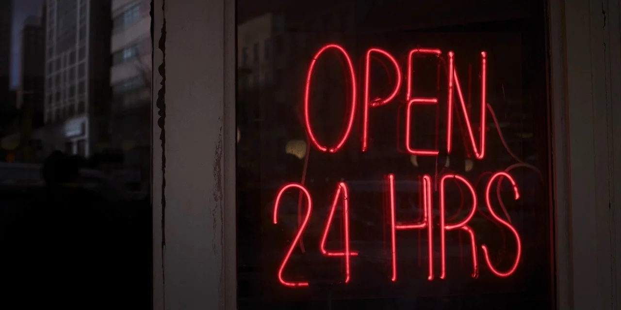 The Sound of Silence movie still: closeup — A glass pane at night displays a bright red neon sign. The sign text reads OPEN 24 HRS against a dark…