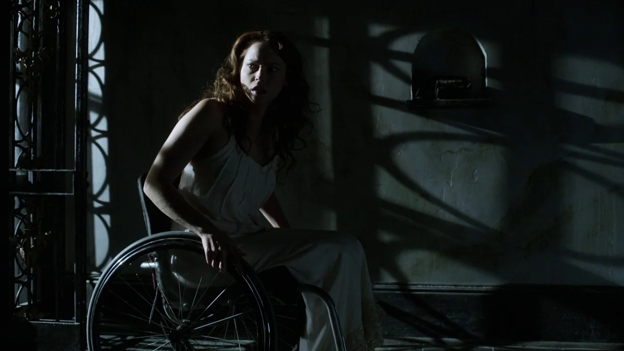 Curse of Chucky (2013) movie still: medium — A dimly lit space features a woman seated in a wheelchair before a wall. Sharp shadows from a nearby…