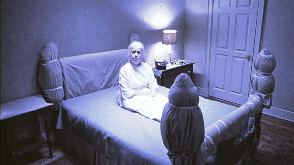 The Exorcist (1973) movie still: medium — An elderly woman sits centered on a bed inside a simple, sparsely furnished room. The wooden bed frame is…