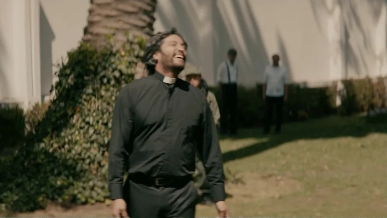 Bel Canto (2018) movie still: medium — A sunny outdoor lawn sits next to a large palm tree and a white wall. A man dressed as a priest smiles while…