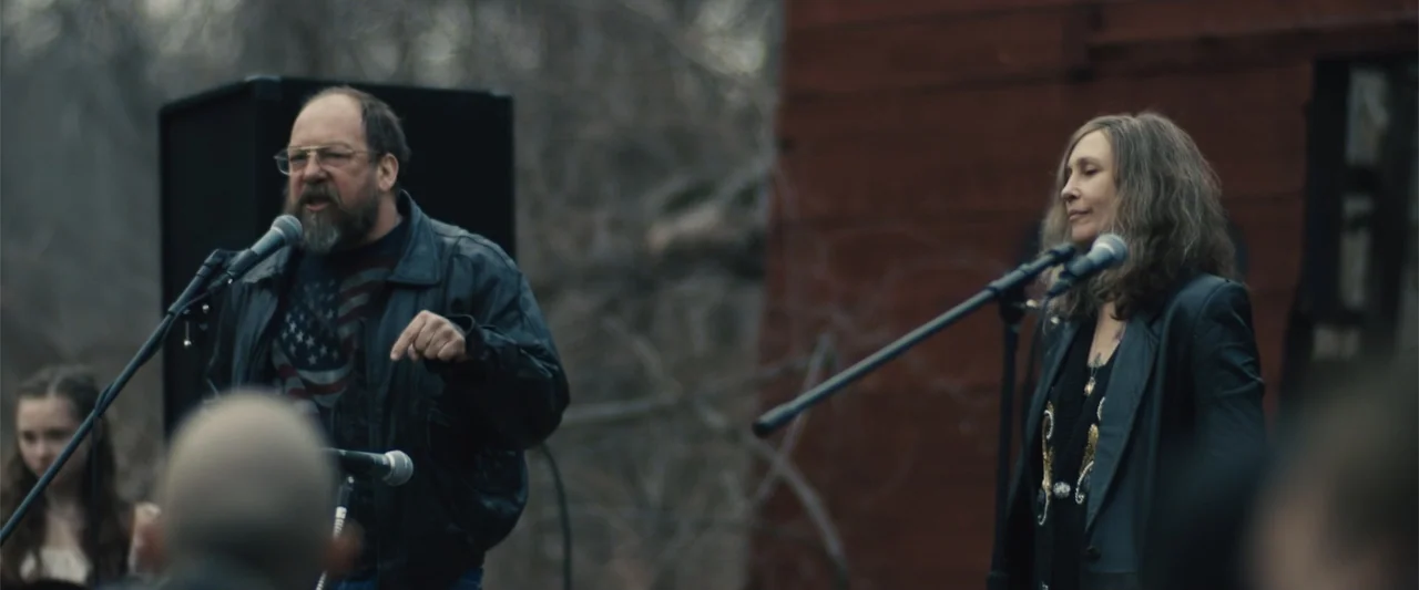 Skin (2009) movie still: medium — An outdoor stage area features a dark red structure in the background and blurred trees. A man with a beard and a…