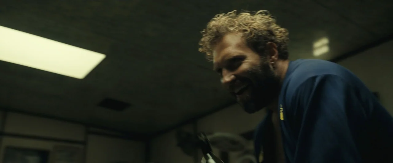 Dangerous Animals (2025) movie still: medium — An interior space with a dark ceiling and drop-panel lighting fixtures. A man with curly blonde hair…