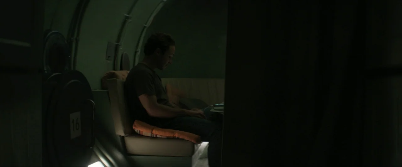 Last Breath (2025) movie still: medium — A dimly lit narrow compartment features curved metal walls and a padded bench seat. A man sits alone on the…