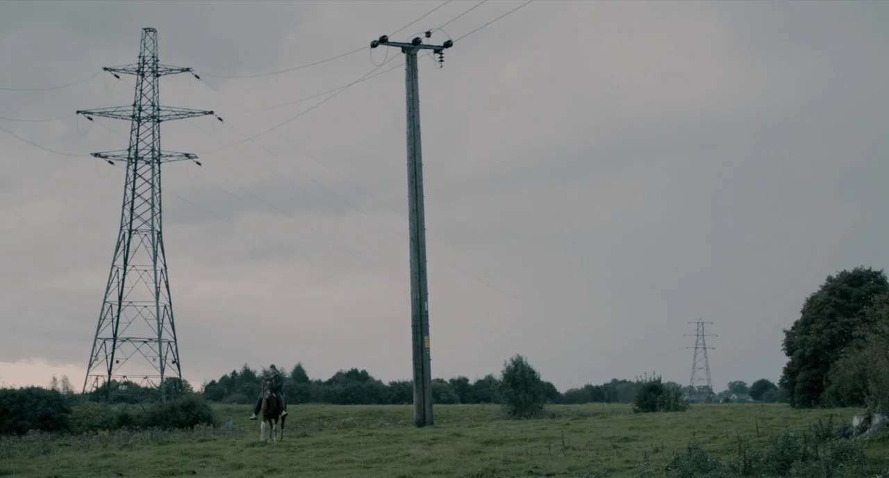 The Selfish Giant (2013) movie still: wide — An expansive open field features large metal transmission towers and a wooden pole. A single rider on…