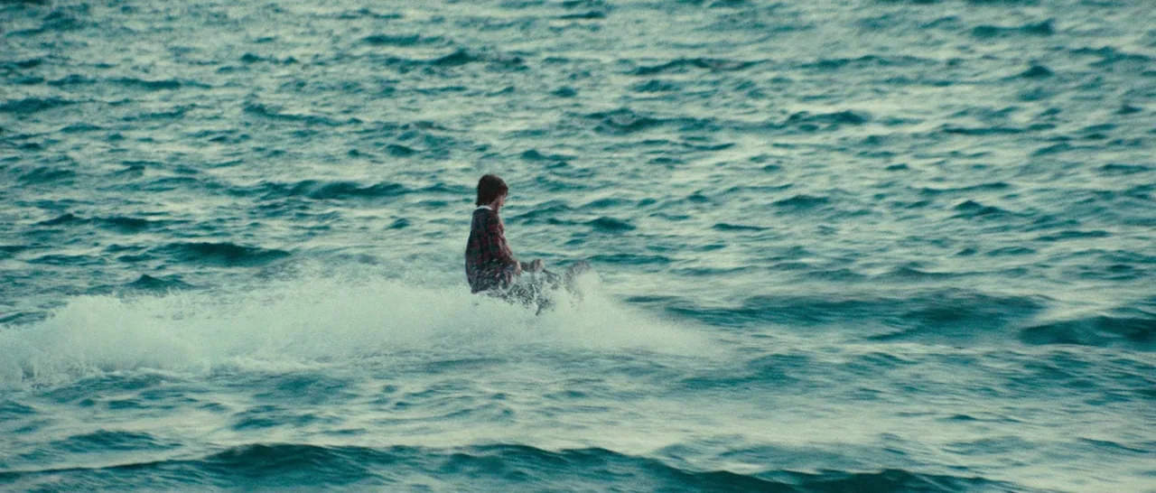 Swiss Army Man (2016) movie still: wide — The open sea displays rhythmic blue-green waves under natural daylight conditions. A single man in a plaid…