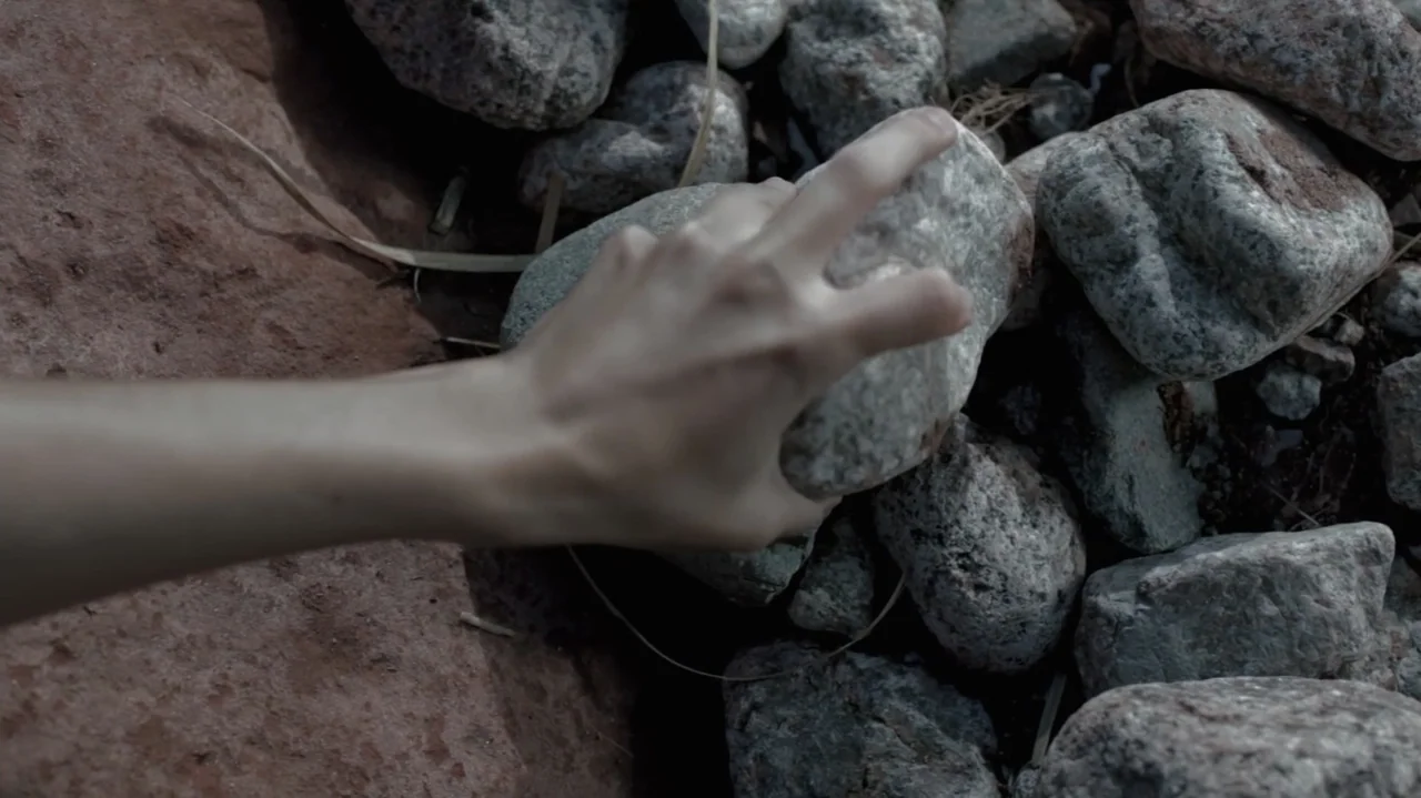 Still Life (2013) movie still: closeup — A pile of grey and brown rocks rests on a dusty reddish surface. A human hand enters the frame from the…