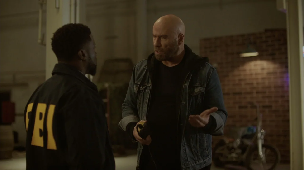 Die Hart (2023) movie still: medium — An interior space features a brick wall, motorcycle, and dim ambient lighting. A bald man in a denim jacket…