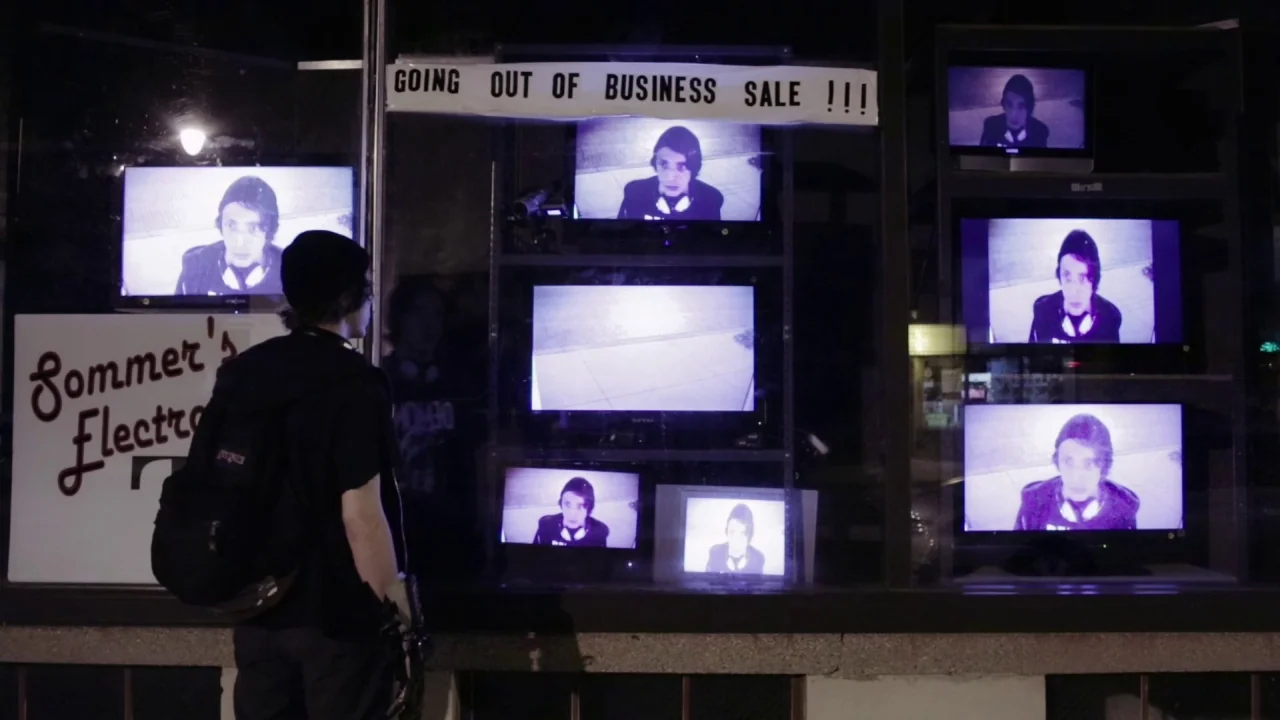 Buzzard (2014) movie still: medium — A nighttime storefront display features several television sets mounted in a cluttered window frame. A teenager…