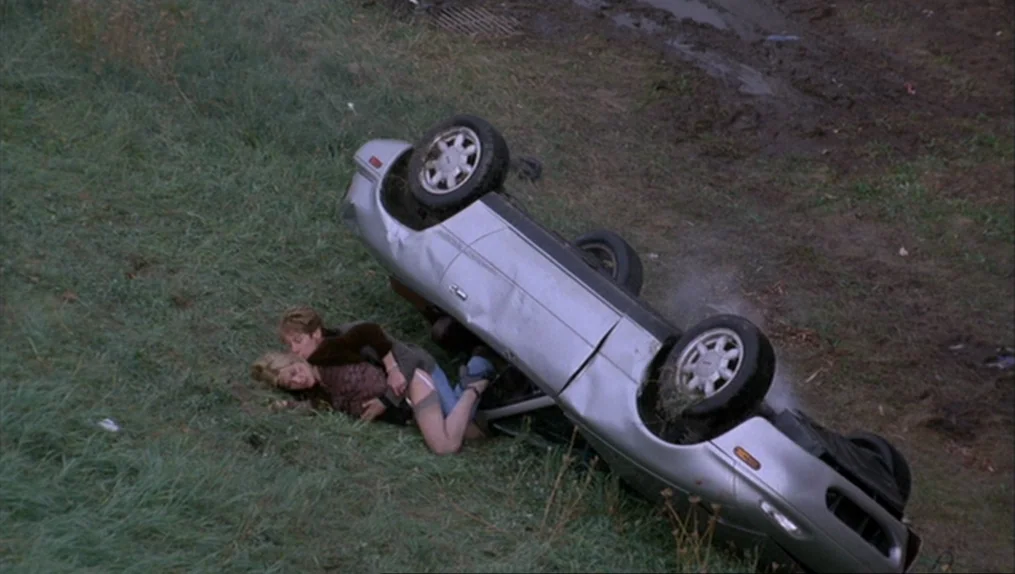 Crash (1996) movie still: wide — An overturned silver sedan rests on a muddy grass embankment near a roadway. Two women are lying on the grass next…