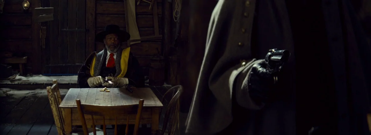 The Hateful Eight (2015) movie still: medium — A dark wooden interior has thick log walls and a small rustic dining table. A man in a hat sits…