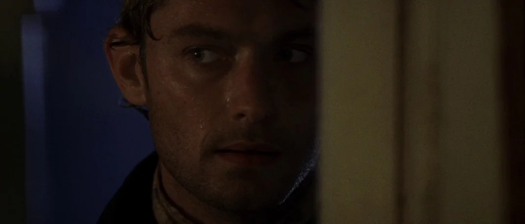 Cold Mountain (2003) movie still: closeup — A man peers around the edge of an indoor wooden door frame. Dim interior lighting obscures parts of his…
