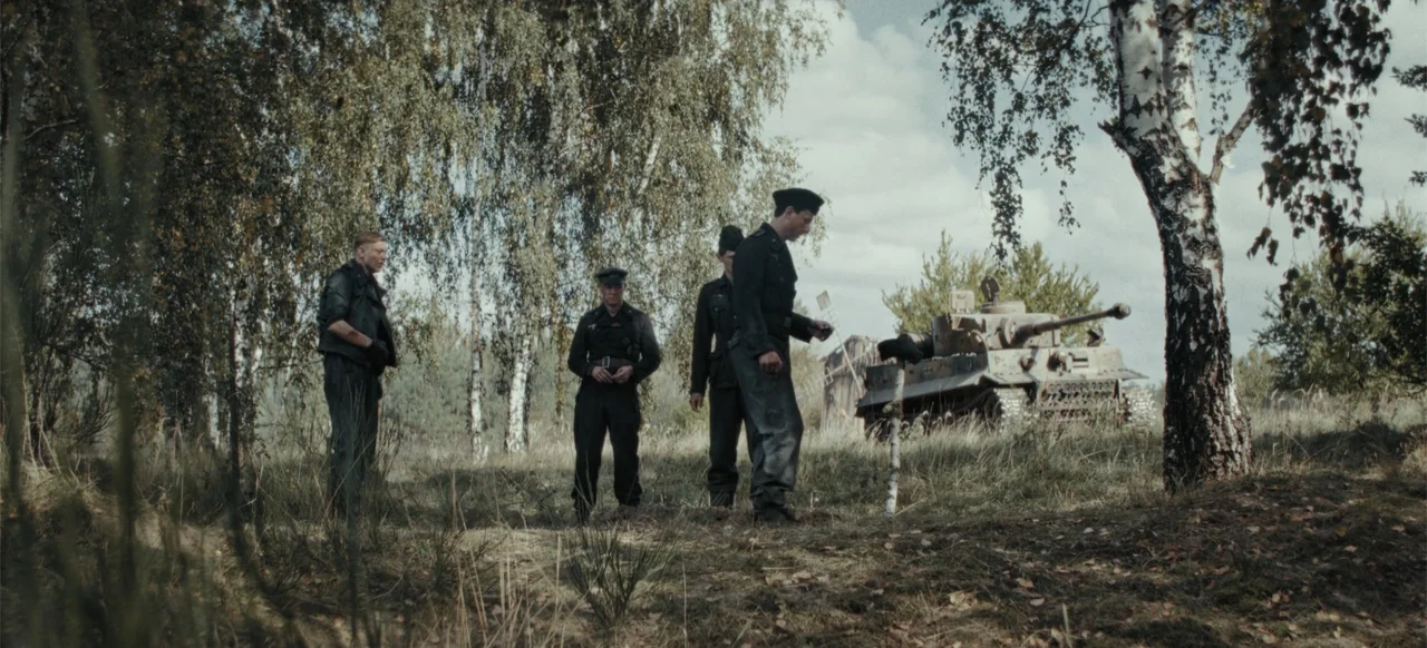 The Tank (2025) movie still: fullbody — A rural landscape with birch trees features a camouflaged tank in the distance. Four soldiers stand together…