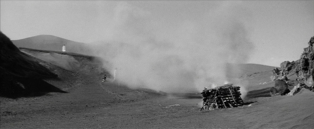 Story of a Prostitute (1965) movie still: wide — A stark volcanic field features a large, smoking wood pyre in the center foreground. In the distant…