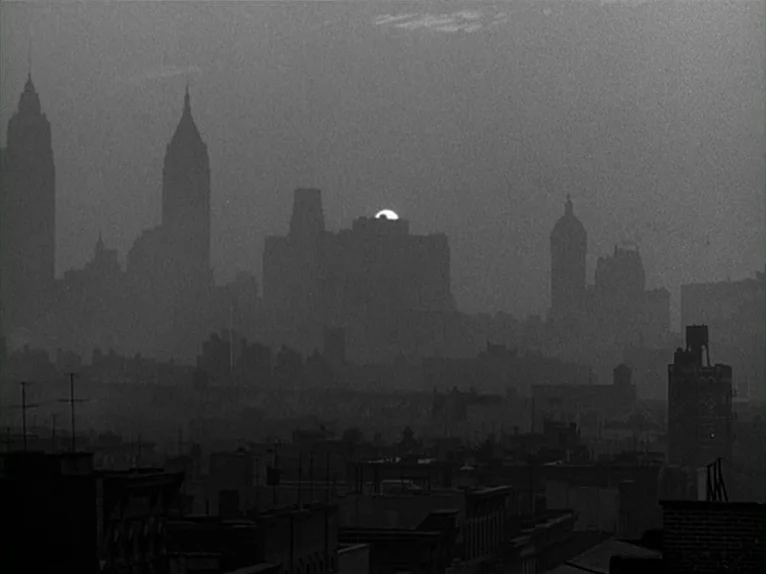 Killer’s Kiss (1955) movie still: establishing — A dark urban landscape features layered city buildings silhouetted against a fading sky. The sun is…