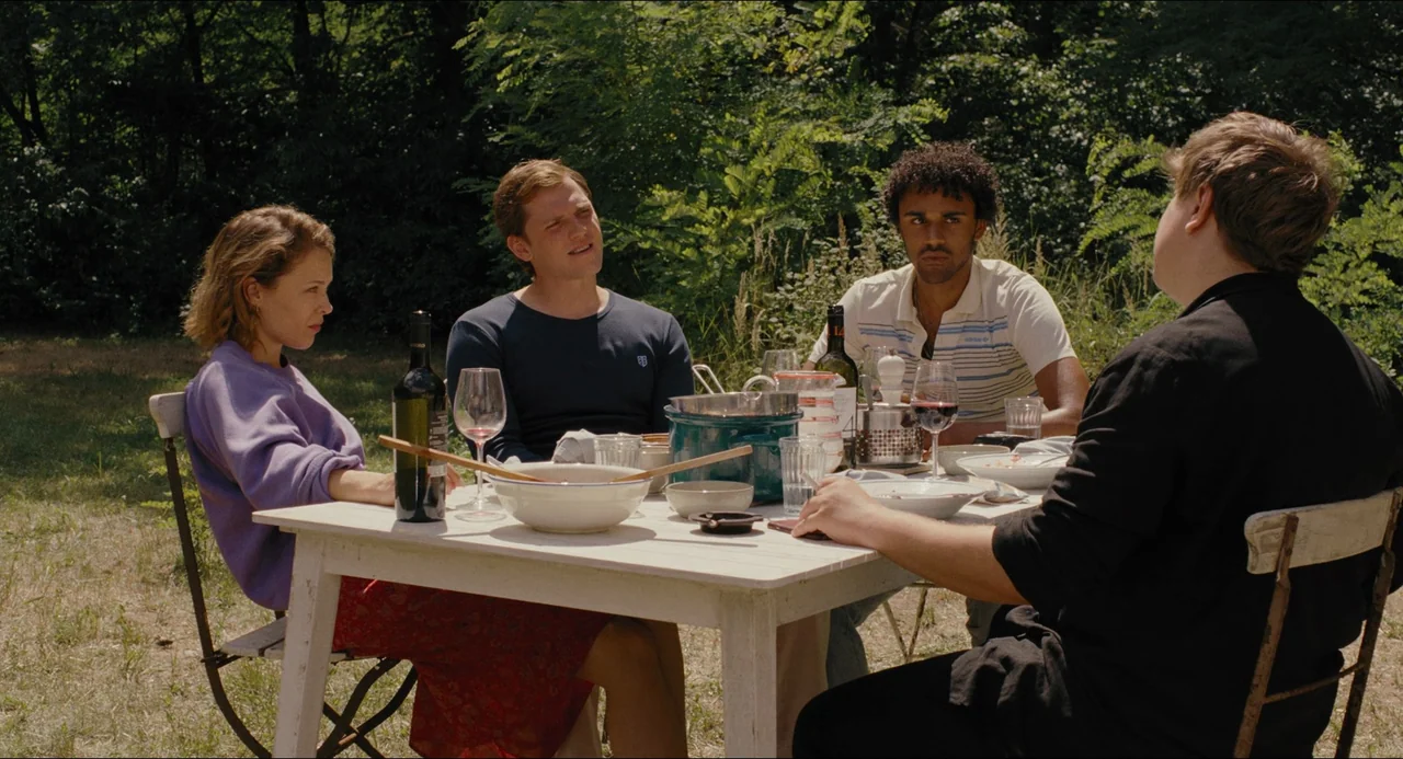 Afire (2023) movie still: medium — Four friends are gathered around a wooden table in a shaded outdoor setting. They are sitting together, eating…