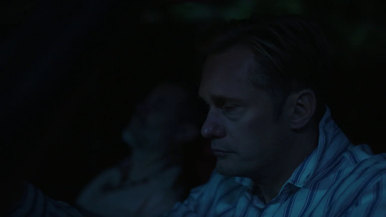 Infinity Pool (2023) movie still: closeup — A dark car interior at night with a man in the driver seat. The man stares ahead while another person is…