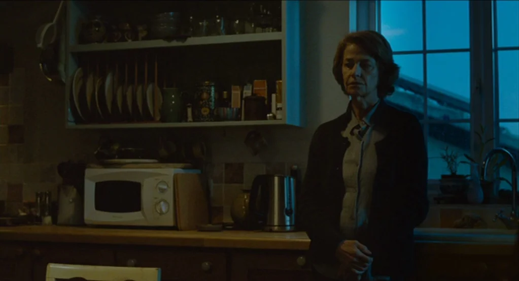 45 Years (2015) movie still: medium — A dark, dimly lit domestic area with wooden countertops and white appliances. A woman stands motionless before…