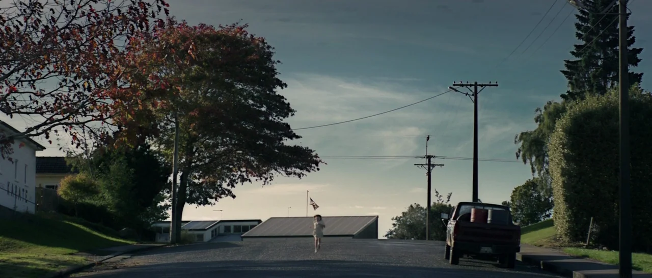Pete’s Dragon (2016) movie still: wide — A suburban street slopes downward with a parked red pickup truck on the right. A single small child runs…