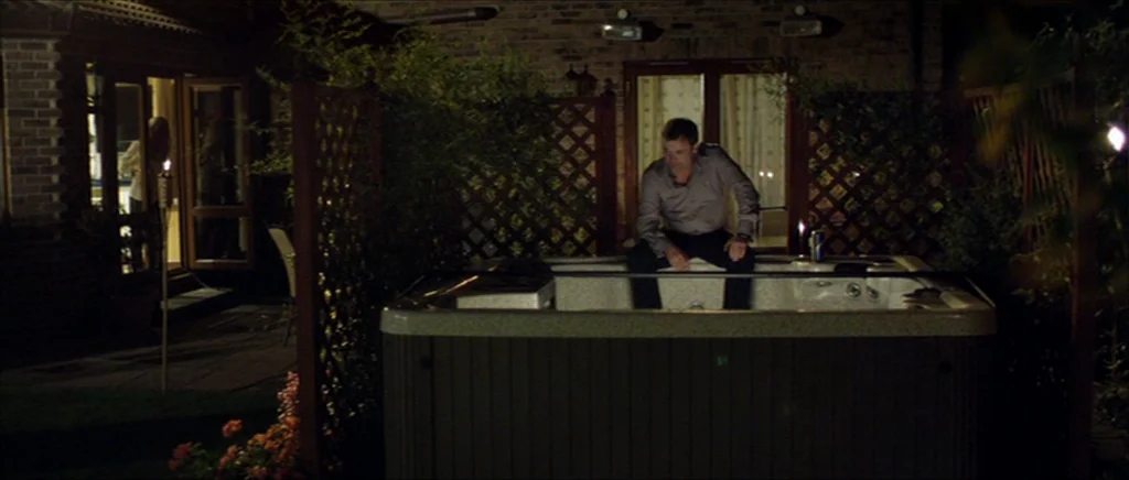 Kill List movie still: wide — A dark outdoor space features a hot tub against a brick wall with lattice panels. A man sits on the edge of the tub…