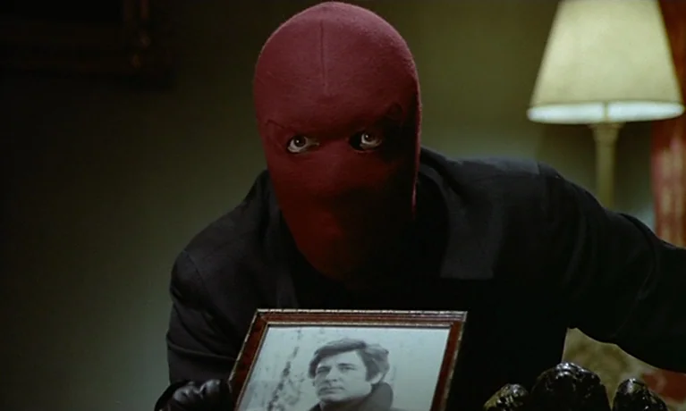 Nuits Rouges (1974) movie still: medium — An interior space with a dimly lit table lamp in the background. A figure wearing a red fabric mask holds…