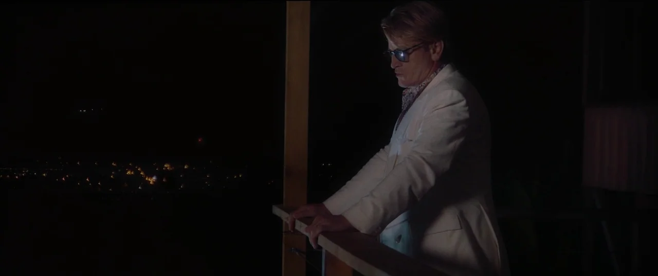 Pacifiction (2022) movie still: medium — A dark, wooden balcony overlooks a distant city at night. A middle-aged man stands at the railing, looking…