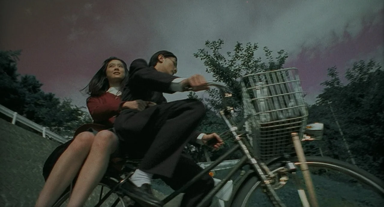 Uzumaki (2000) movie still: medium — A man and woman ride a bicycle up a steep outdoor incline at dusk. The sky has a purple hue while lush green…