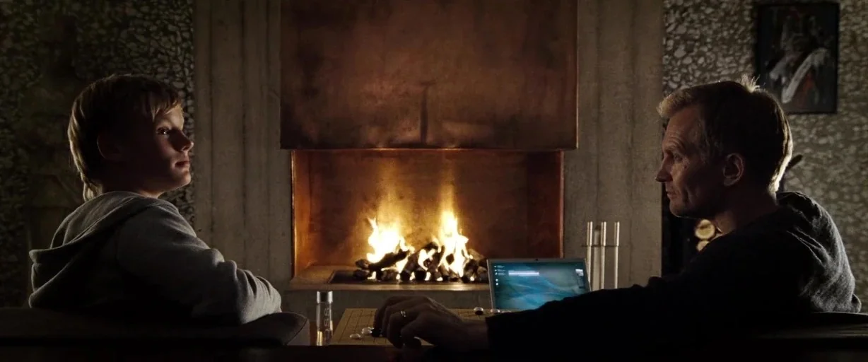 The International (2009) movie still: medium — A modern interior space features a large fireplace with a copper hood facade. A boy and a man sit at…