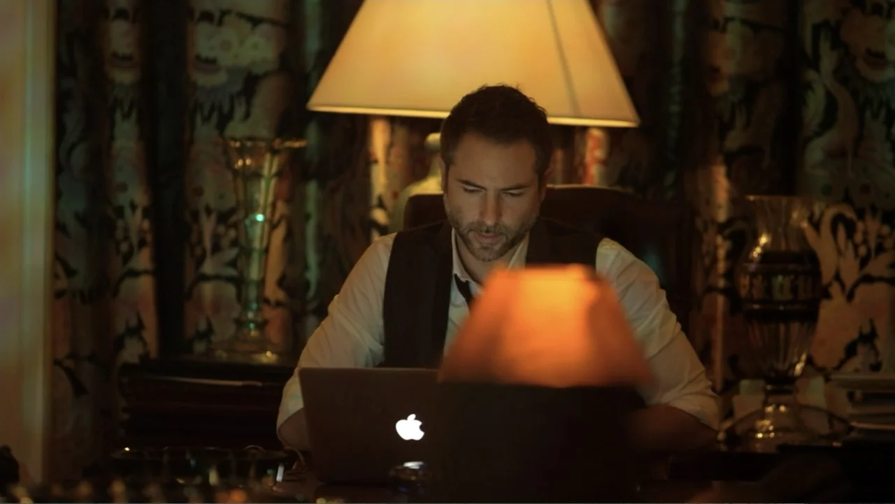 Entre las sombras (2025) movie still: medium — A dimly lit home study features patterned wallpaper and antique decorative glass vases. A man in a…