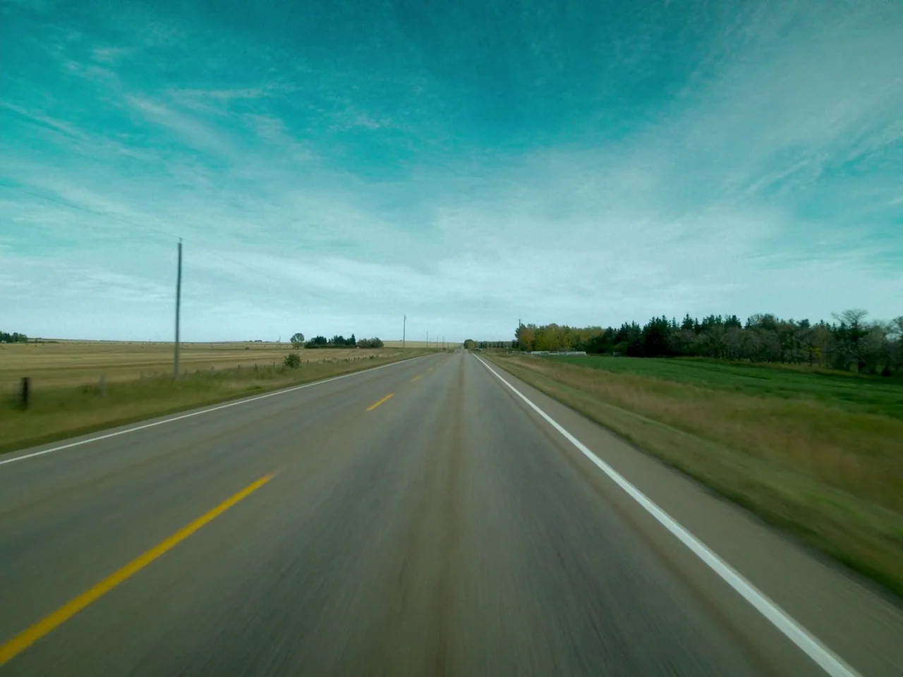 Die My Love (2025) movie still: establishing — A rural highway stretches toward the horizon through open fields and patches of trees. A single paved…