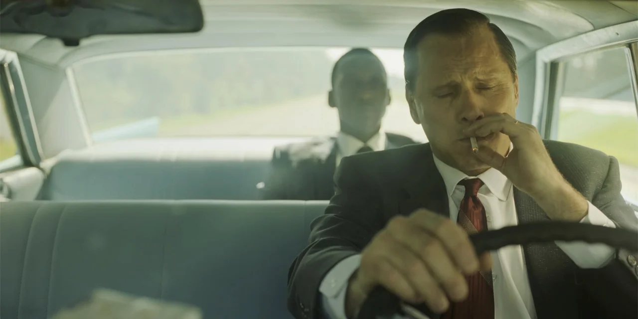 Green Book (2018) movie still: medium — A 1960s sedan interior features light blue bench seats and large windows. The driver in a suit holds a…