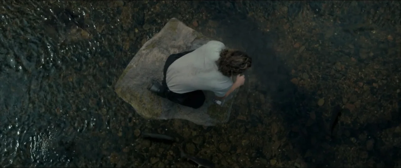 Kraven the Hunter (2024) movie still: medium — A man kneels on a solitary rock amidst shallow flowing water with visible riverbed stones. The man is…